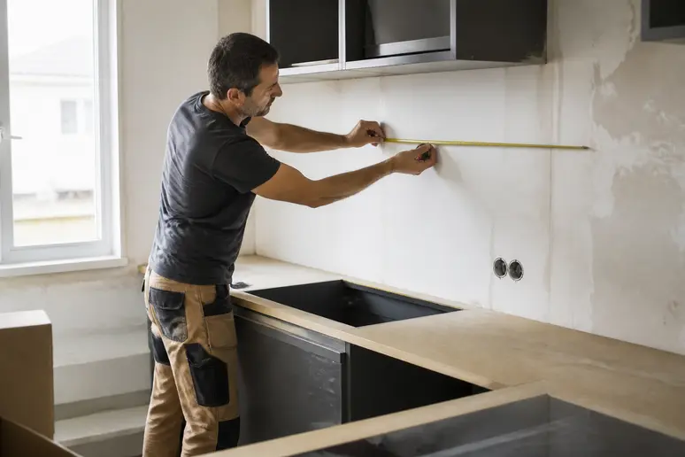 Un artisan mesure une section de mur dans une cuisine en cours de rénovation, soulignant le budget nécessaire pour les travaux.