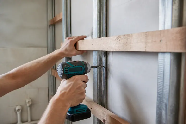 Mains installant des tasseaux en bois entre des montants métalliques dans un mur en placo pour renfort.
