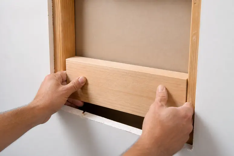 Main droite insérant une planche de bois dans une ouverture derrière une plaque de plâtre.
