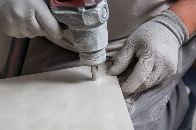 Mains d'artisan amorçant un trou dans un carreau de grès cérame avec une scie cloche diamantée en biseau.
